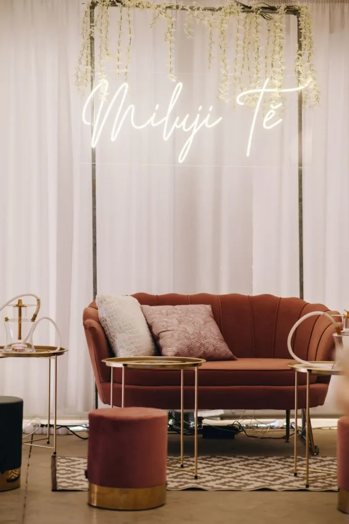 Stylový svatební fotokoutek a lounge s neonovým nápisem 'Miluji Tě', terakotovou pohovkou, květinami a vodními dýmkami na zlatých stolcích.