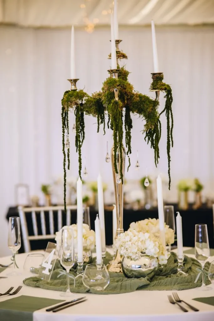 Elegantní svatební výzdoba kulatého stolu s vysokým stříbrným svícnem, mechem a bílými květy pro slavnostní hostinu.